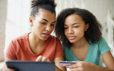 Two young women using a tablet and credit card while shopping online indoors.