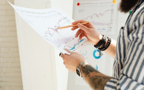 A man analyzing stock market charts with a pen, holding a paper report indoors.