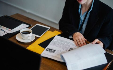 High angle of crop faceless businesswoman in formal clothes sitting at table with tablet and hot coffee and looking through documents