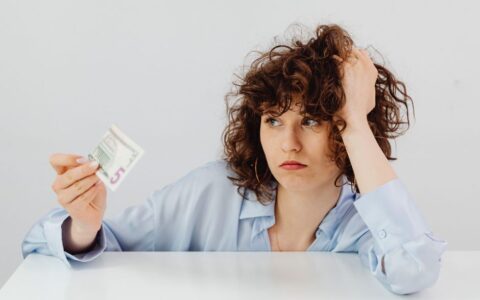 Concerned woman with curly hair holding a five dollar bill, expressing financial worry.