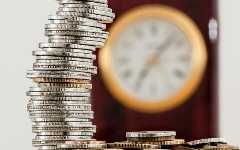 A close-up image of stacked coins with a blurred clock, symbolizing time and money relationship.