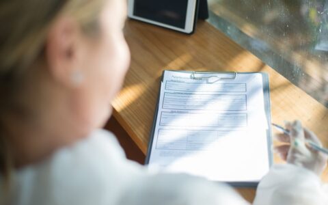 A woman fills out an application form with a pen at a sunlit wooden desk.